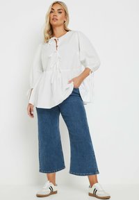 Witte peplumblouse met pofmouwen en strikdetails, gecombineerd met donkerblauwe wijde jeans en witte sneakers met zwarte strepen.