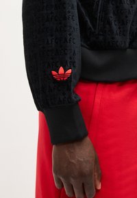 Tume nahaga käsi, mis kannab musta dressipluusi punase Adidas logo ja punaste pükstega, nähtav randmest vööni.