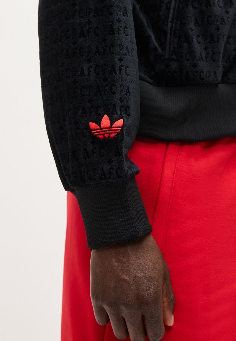 Donkere hand met zwarte sweatshirt met rood Adidas-logo en rode broek, zichtbaar van pols tot taille.
