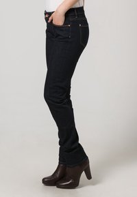 Jeans blu scuro con vestibilità slim, cinque tasche, cuciture dorate e accessori in metallo. Indossati con stivali marroni con tacco alto.