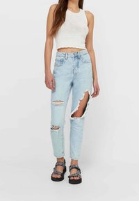 Lichtblauwe ripped jeans met rafelige randen, hoge taille en grote uitsnijdingen op de benen. Gecombineerd met een witte crop top en zwarte sandalen.