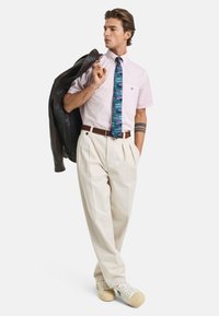 Lichtroze shirt met korte mouwen en een borstzak, blauwe gestreepte stropdas, witte geplooide broek, een leren bruine riem, en een bruine leren jas over één schouder gedrapeerd.