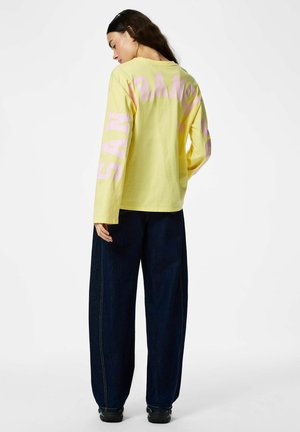 Femme de dos portant un t-shirt jaune à manches longues avec le texte rose "SAN FRANCISCO" et un pantalon ample bleu foncé, sur fond blanc.