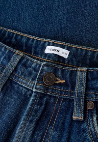 Jeans hlače iz jeansa z modrim pranjem, z bakreno sponko, kontrastnim šivom in etiketo z napisom "TWO SOON 31/32" na pasu.