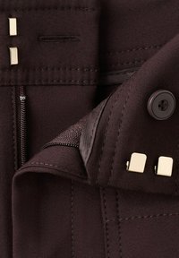 Prim-plan cu pantaloni din material închis la culoare care se încheie cu un nasture, fermoar și două cârlige metalice pe talie.