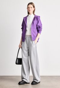 Fialová mikina na zips, biele priliehavé tričko, šedé široké tepláky s logom, čierna taška a masívne čierne topánky na neutrálnom pozadí.