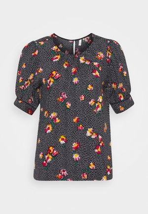 Blouse noire à petits pois blancs, motif floral coloré, manches courtes bouffantes et encolure ronde avec fermeture boutonnée au dos.