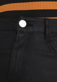 Svarta denimbyxor med en slät yta, iögonfallande silverfärgad metallknapp med ett ingraverat logotyp och kontrasterande sömmar.