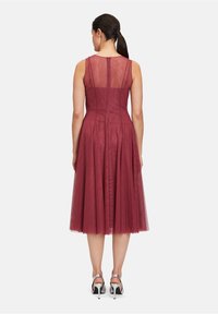 Weinrotes Kleid mit tailliertem Oberteil, Tüllrock und durchsichtiger Rückenpartie. Mit einem Reißverschlussverschluss und silbernen High-Heels.