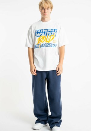 Młoda blond osoba stojąca, ubrana w biały, za duży T-shirt z niebieskim i żółtym napisem oraz luźne, ciemnoniebieskie spodnie i białe buty.