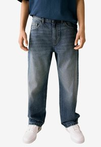 Jean en denim bleu clair délavé avec une coupe décontractée, comportant cinq poches et des coutures contrastantes, associés à des baskets blanches.