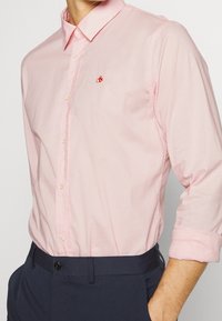 Camicia button-up rosa chiaro con colletto classico e maniche arrotolate. Presenta un piccolo logo rosso sul lato sinistro del petto. Tessuto in cotone texturizzato.