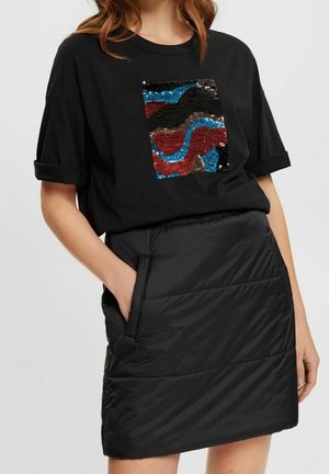 Kobieta w czarnej koszulce z wielokolorowym, cekinowym, abstrakcyjnym kwadratem oraz czarnej pikowanej spódnicy z kieszeniami, z ręką w kieszeni.