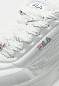 White sneaker with textured mesh upper, flat laces, and rubber sole. Features FILA logo in blue and red on side. Smooth, shiny accents.