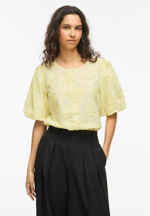 Femme aux cheveux bouclés et foncés portant un chemisier jaune pâle à manches bouffantes avec broderies florales et un pantalon plissé noir taille haute.