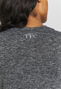 Camiseta deportiva gris jaspeada con cuello redondo, que presenta un pequeño logo reflectante en la parte posterior. El material parece tener textura y ser ligero.