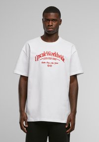 Man wearing white oversized T-shirt with red text "Upscale Worldwide" and cities listed, standing against plain gray background.