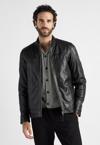 Svart läderjacka med ståkrage och vadderade axeldetaljer, kombinerad med en randig henleytröja och mörka byxor.