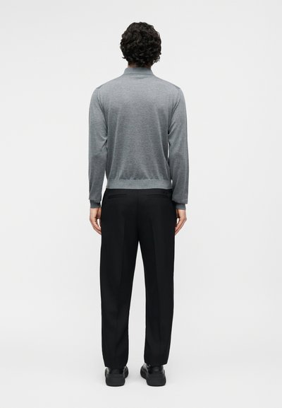 Homme aux cheveux bouclés portant une chemise grise à manches longues, un pantalon noir et des chaussures noires, debout de dos sur un fond uni.