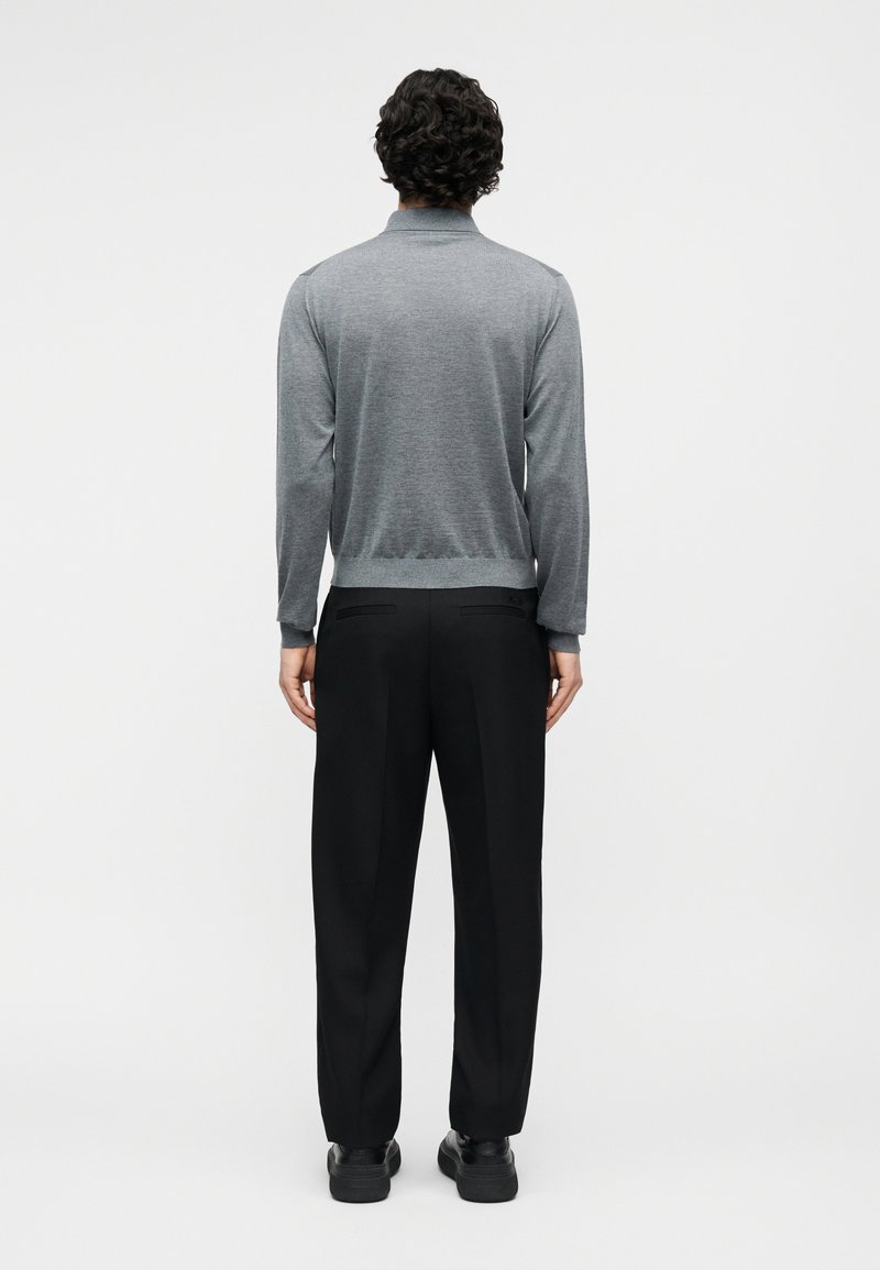 Man met krullend haar, gekleed in een grijs shirt met lange mouwen, zwarte broek en zwarte schoenen, staand met de rug naar de camera tegen een effen achtergrond.