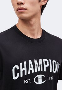 Νεαρός άνδρας που φοράει μαύρο T-shirt με λευκό κείμενο και λογότυπο "CHAMPION EST. 1919", κοιτάζοντας ελαφρώς στο πλάι σε απλό φόντο.