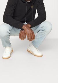 Musta huppari kultaisella logokuvioinnilla, vaaleansiniset farkut ja valkoiset tennarit kuminanaisella pohjalla. Rentoon tyyliin kuuluva asukokonaisuus, jossa on väljää istuvuutta ja tekstuurien kontrastia.