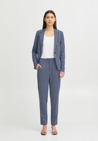 Un costume à carreaux bleu marine avec un blazer ajusté et un pantalon fuselé, fabriqué dans un tissu lisse, complété par un haut blanc et des talons noirs.