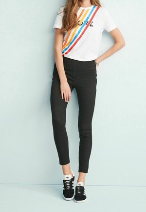 Femme portant un T-shirt blanc avec des rayures colorées et du texte, un pantalon skinny noir et des baskets Adidas noires, debout devant un mur bleu clair.