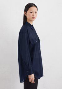 Marineblauwe, langemouwen shirt van doorzichtig materiaal met een knoopsluiting aan de voorkant en twee borstzakken. Kenmerkt zich door manchetten en een klassieke kraag.
