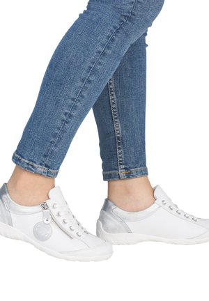 Personne portant un jean bleu et des baskets blanches décontractées avec des accents argentés et une fermeture éclair latérale, debout sur un fond blanc.