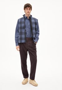 Herrenoutfit mit einer blauen karierten Weste über einem passenden Hemd, kombiniert mit dunklen Cordhosen und hellen Slippern.