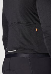 Czarna, sportowa bluzka z długim rękawem z zamkiem błyskawicznym, gładka w dotyku, z odblaskowymi detalami. Dopasowany krój w połączeniu z czarnymi legginsami.