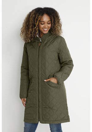 Donna sorridente, che indossa un cappotto trapuntato verde oliva fino al ginocchio con cerniera e mano in tasca, in piedi davanti a uno sfondo chiaro.