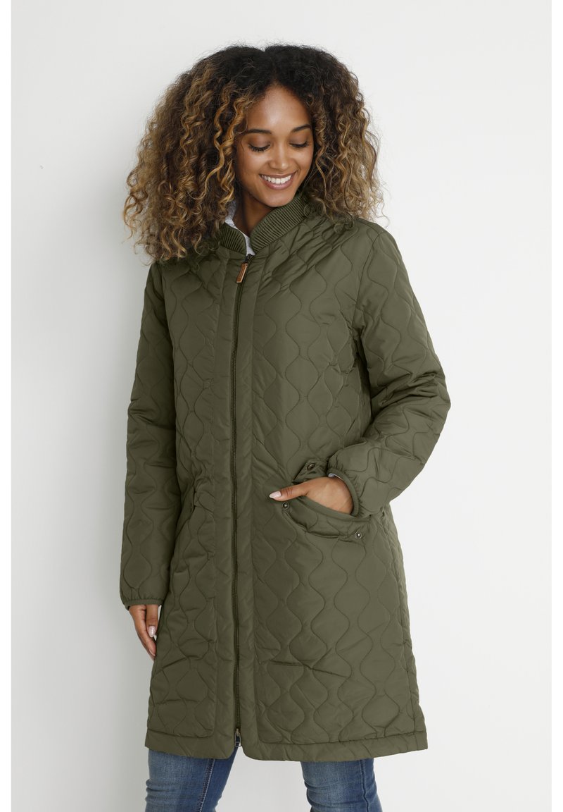 Nainen hymyilee, pukeutuneena polvenmittaiseen oliivinvihreään tikattuun takkiin, jossa on vetoketju ja käsi taskussa, seisoo vaaleaa taustaa vasten.