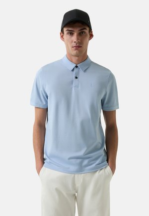 Hellblaues Poloshirt mit klassischem Kragen, Drei-Knopf-Leiste und kleinem Logo auf der Brust. Kombiniert mit weißen Hosen.