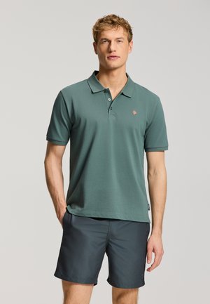 Muž stojaci v zelenej krátkosleeve polo košeli a tmavosivých šortkách, ruky vreckách, na jednofarebnom pozadí.