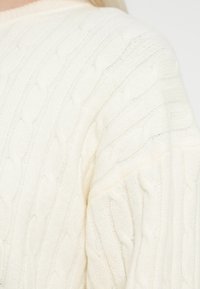 Pulover tricotat de culoare crem, cu model de cablu, având un decolteu rotund și umeri căzuți, evidențiind un material moale și texturat.