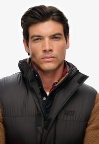 Ung man med mörkt hår iförd en brun fleece och en svart puffer-väst med "Superdry"-logotyp, som tittar direkt in i kameran.