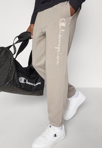 Ανοιχτό γκρι φόρμα με ελαστικά μανσέτα, με εμφανές κεντητό λογότυπο "Champion" στο πλάι. Συνδυασμένο με μαύρη τσάντα duffel και λευκά αθλητικά παπούτσια.