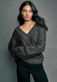 Grå strikket sweater med V-hals og løse ærmer, der har et struktureret mønster og en subtil glans. Båret med mørke bukser, foran en grå baggrund.