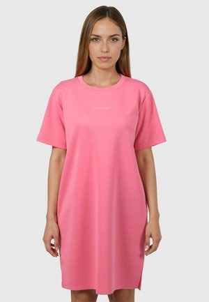 Kvinde står op af en ensfarvet baggrund iført en løstsiddende, knælængde lyserød kjole med korte ærmer og små hvide bogstaver på brystet.