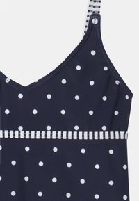 Marineblauer Badeanzug mit weißen Punkten, verstellbaren Trägern und einem gestreiften dekorativen Band an der Taille. Glatte Stoffstruktur.