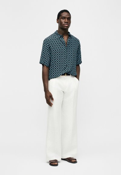 Άνδρας στέκεται φορώντας κοντομάνικο πουκάμισο σε ναυτικό μπλε με ανοιχτό μπλε μοτίβο, λευκό παντελόνι φαρδύ και σκούρα σανδάλια, μπροστά σε απλό φόντο.