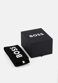 Caja negra con el logo blanco de "BOSS", junto a un estuche negro a juego con el logo. El estuche tiene un acento a rayas y una textura suave.