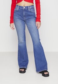 Jeans de denim azul acampanados con cintura alta, diseño clásico de cinco bolsillos y un ligero desgaste. Combinados con sandalias de plataforma negras.