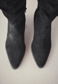 Paar schwarze Wildleder-Stiefeletten mit spitzer Zehenkappe und Reißverschlüssen an der Innenseite, getragen zu schwarzen Hosen auf hellem Boden.