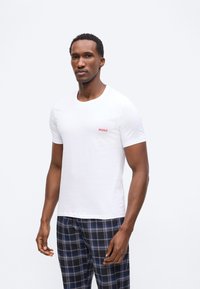 Άνδρας που φοράει λευκό κοντομάνικο μπλουζάκι με κόκκινο λογότυπο "HUGO" και μπλε-μαύρο καρό παντελόνι, στέκεται μπροστά σε απλό λευκό φόντο.