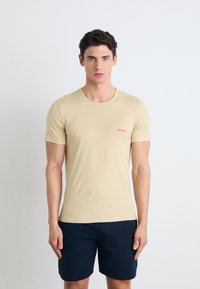 Beige tettskjorte med kort erm, rund hals og liten rød "HUGO" logo på brystet. Brukt sammen med mørk marineblå shorts.