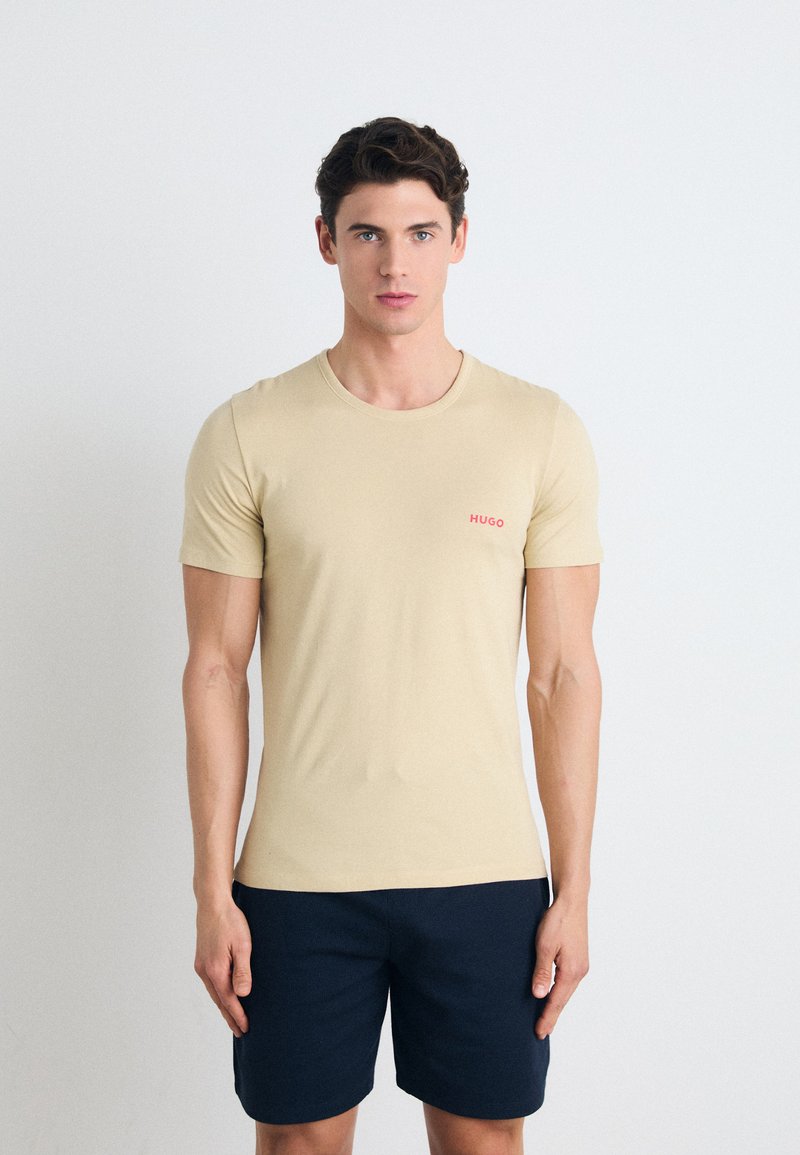 Beige tettskjorte med kort erm, rund hals og liten rød "HUGO" logo på brystet. Brukt sammen med mørk marineblå shorts.
