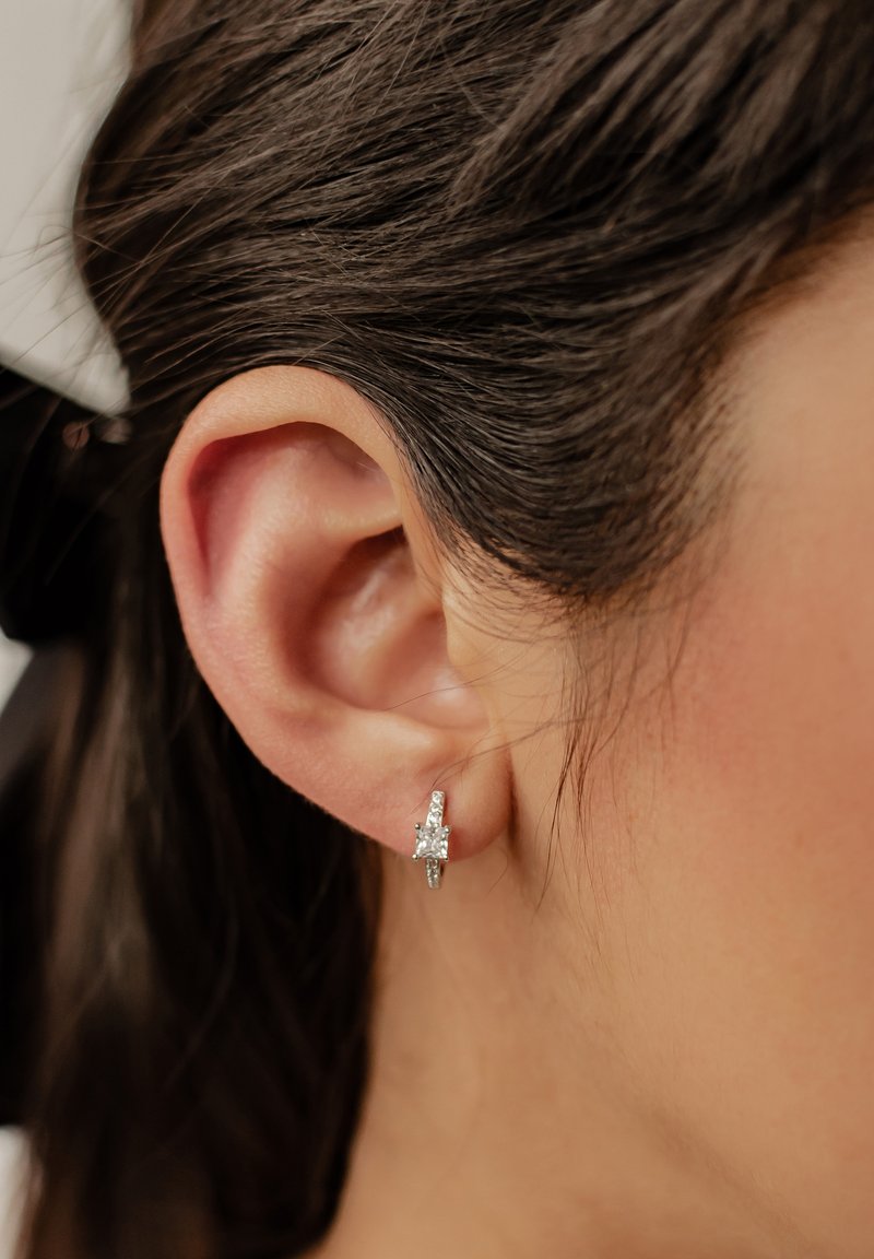 Boucle d'oreille en argent avec une pierre précieuse taillée en carré et transparente, présentant des détails complexes et une finition polie, portée sur l'oreille d'une personne.